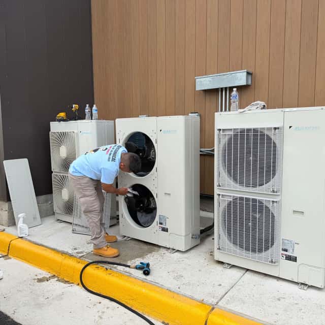 Technician crouched beside a mini-split condenser, cleaning its coil