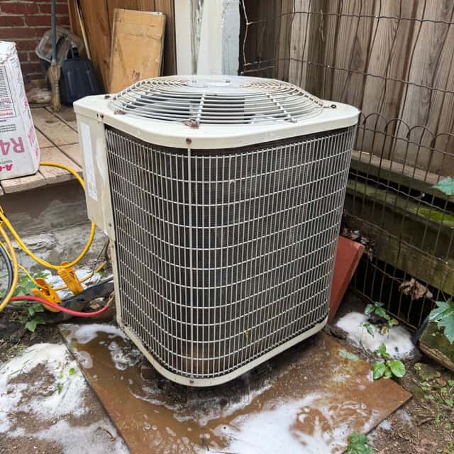 Residential condenser beside a brick wall, its coil fouled with cottonwood fluff