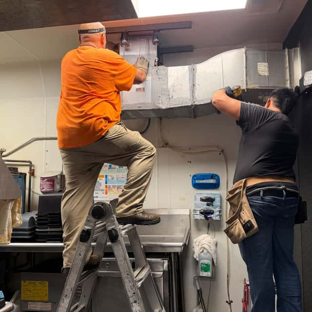 Two-person crew installing sheet-metal ductwork above a commercial kitchen